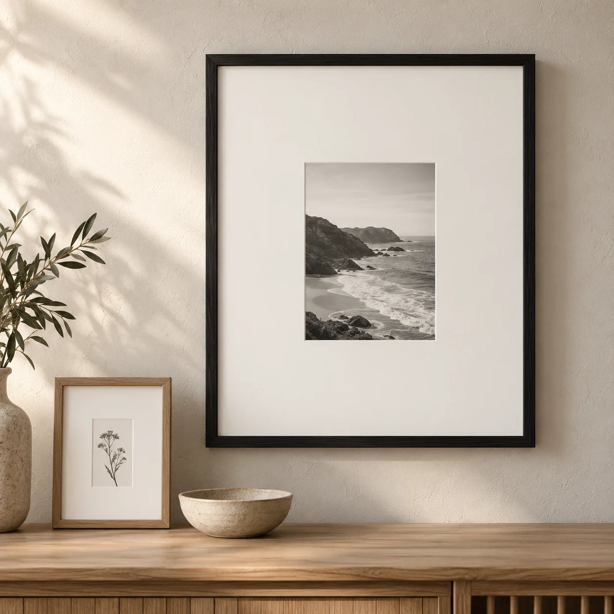 Small monochrome coastal photograph in a black frame with an oversized white mount, beside a mounted botanical drawing