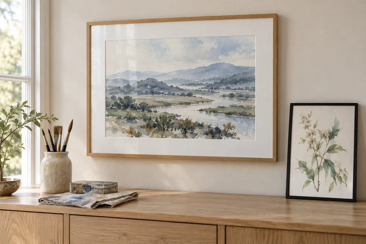 A watercolour river landscape in an oak frame with a white mount, beside a botanical watercolour in a slim black frame